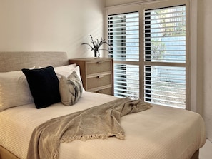 Un dormitorio con cama, almohadas, una mesita de noche con un florero y una ventana con persianas.