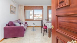 A room with a purple sofa, a small dining table with chairs, a window with curtains, and a wooden door. - Apartment in Roses near Nova Sandy Beach (Rosas / Roses)