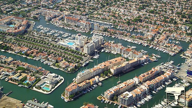 Vista aérea de un puerto deportivo con numerosas embarcaciones amarradas, rodeado de edificios residenciales y una gran estructura hotelera.
