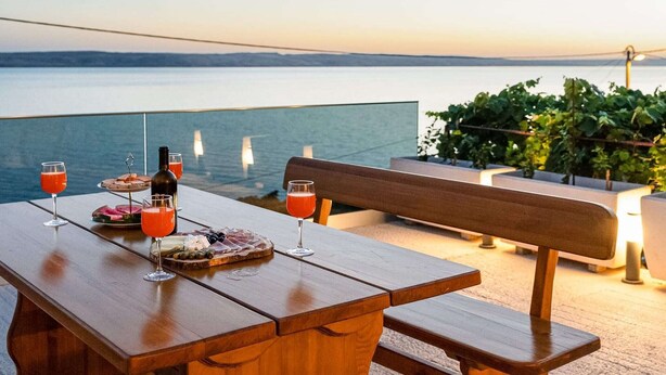 A wooden table with a bench, glasses of red drink, and a bottle on a terrace overlooking a body of water.