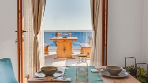 A dining area with a table set for two, a view of the sea, and a wooden table with a bottle and bowls.