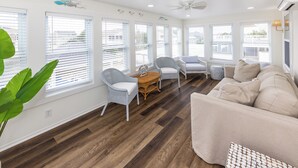 Amplia sala de estar con un sofá beige grande, dos sillones, una mesa de centro y una mesa de comedor. El ambiente tiene piso de madera, paredes blancas y ventanas amplias con persianas.