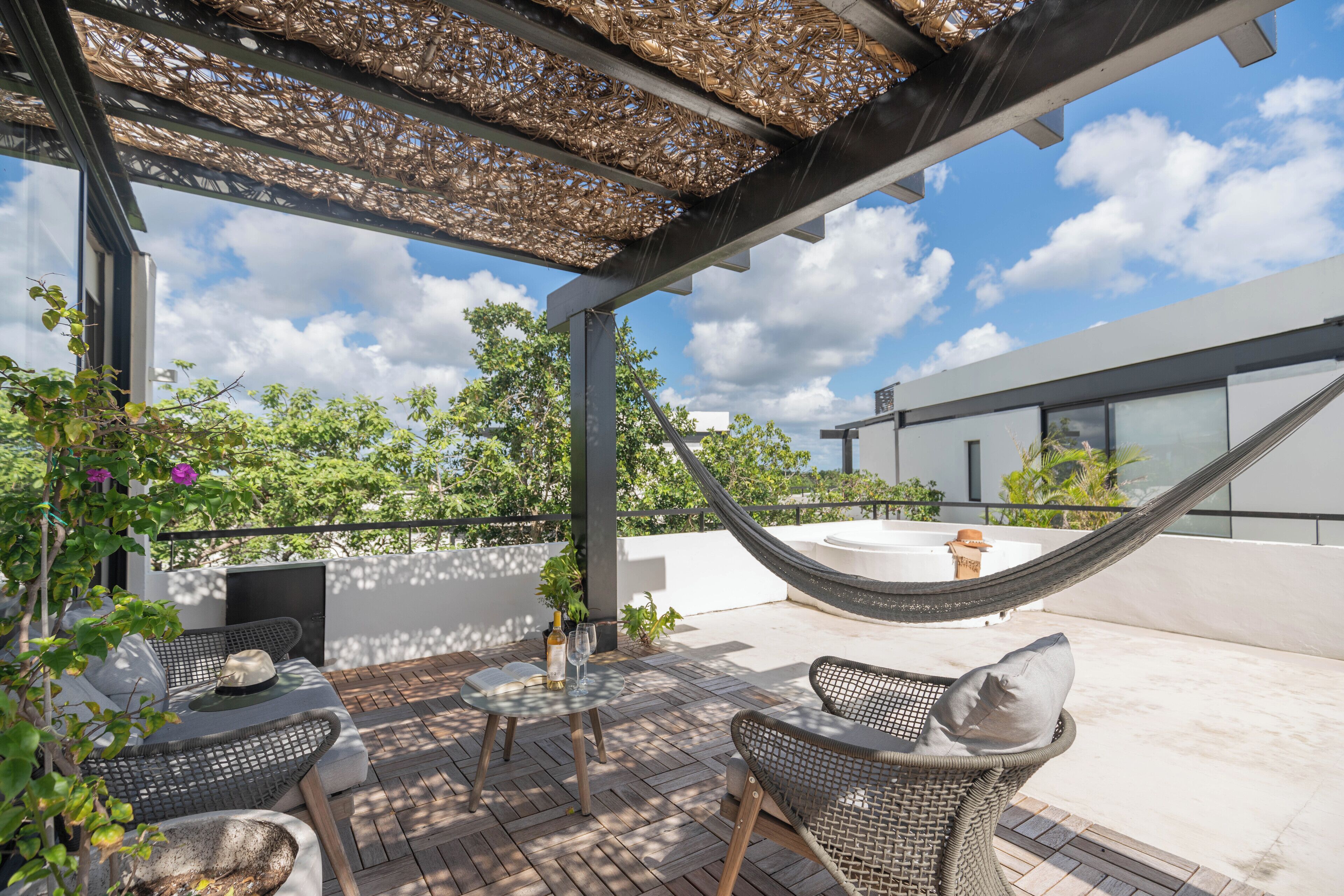 Un patio en la azotea con mobiliario de mimbre, una hamaca y vistas a árboles y un edificio.