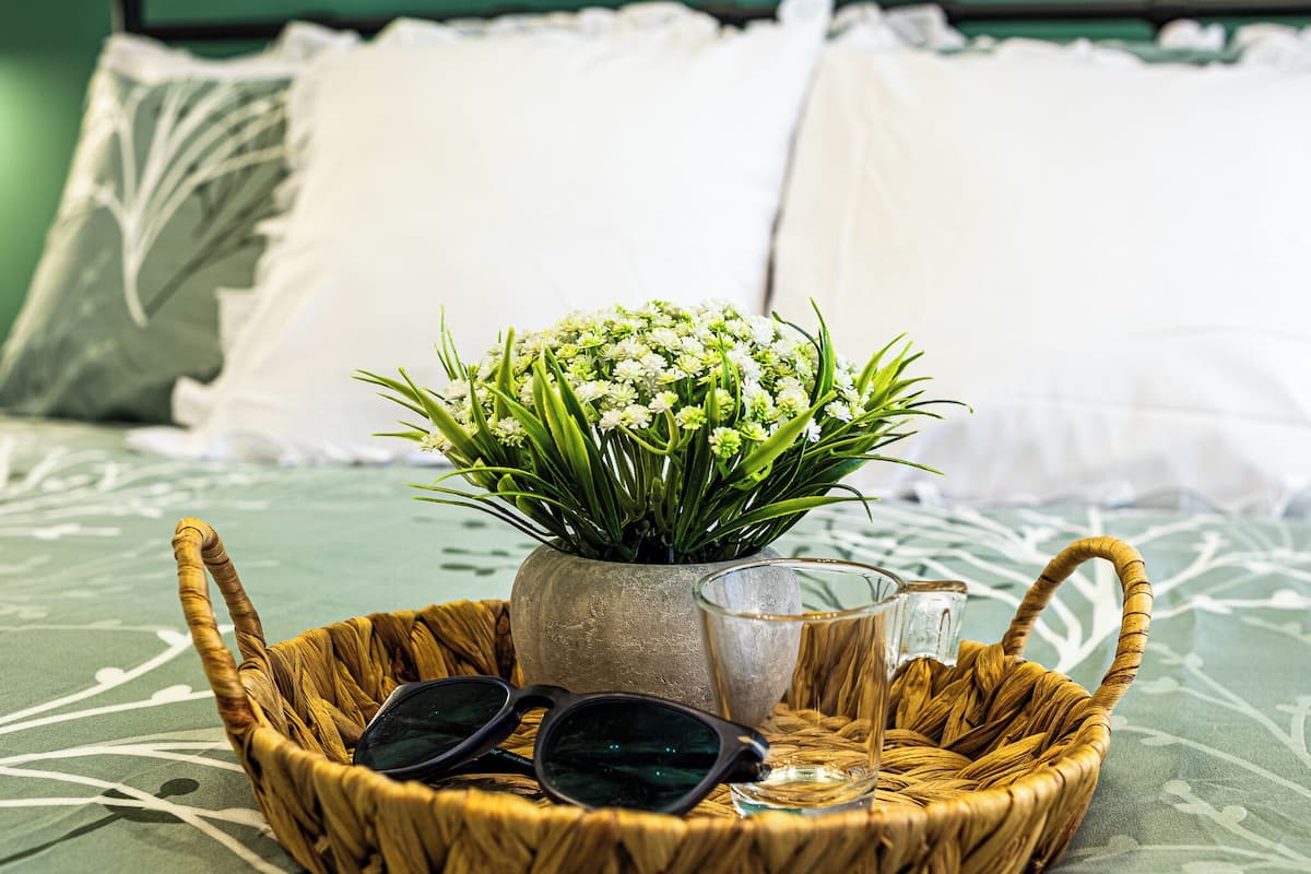 A wicker basket on a bed with a green patterned bedspread, containing a vase with white flowers, a pair of sunglasses, and a glass.