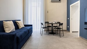 A compact living space with a dark blue sofa, a dining table with four chairs, and a white air conditioning unit on the wall. - Alyde Apartments (Naples)