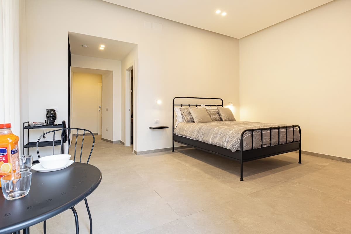 A bedroom with a bed, a table with a chair, and a kitchenette area.