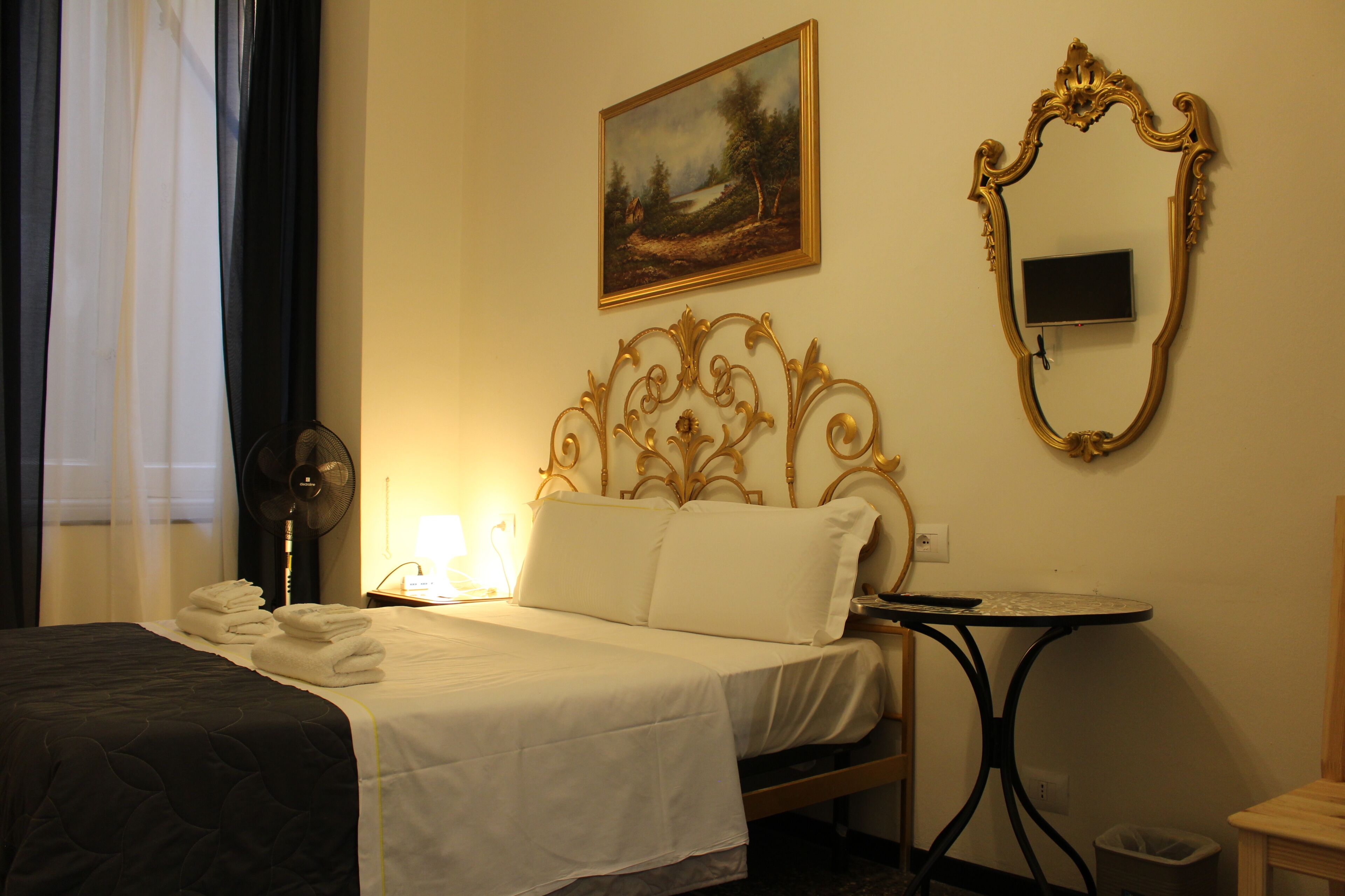 Habitación de hotel con cama, mesita de noche, escritorio, silla, ventilador, un cuadro y un espejo.
