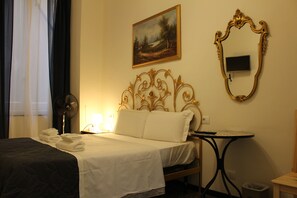 Habitación de hotel con cama, mesita de noche, escritorio, silla, ventilador, un cuadro y un espejo.