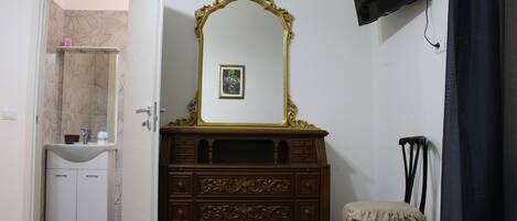 Una habitación con un aparador de madera, un espejo, un pequeño cuadro enmarco, un televisor montado en la pared y una puerta que da a un baño.