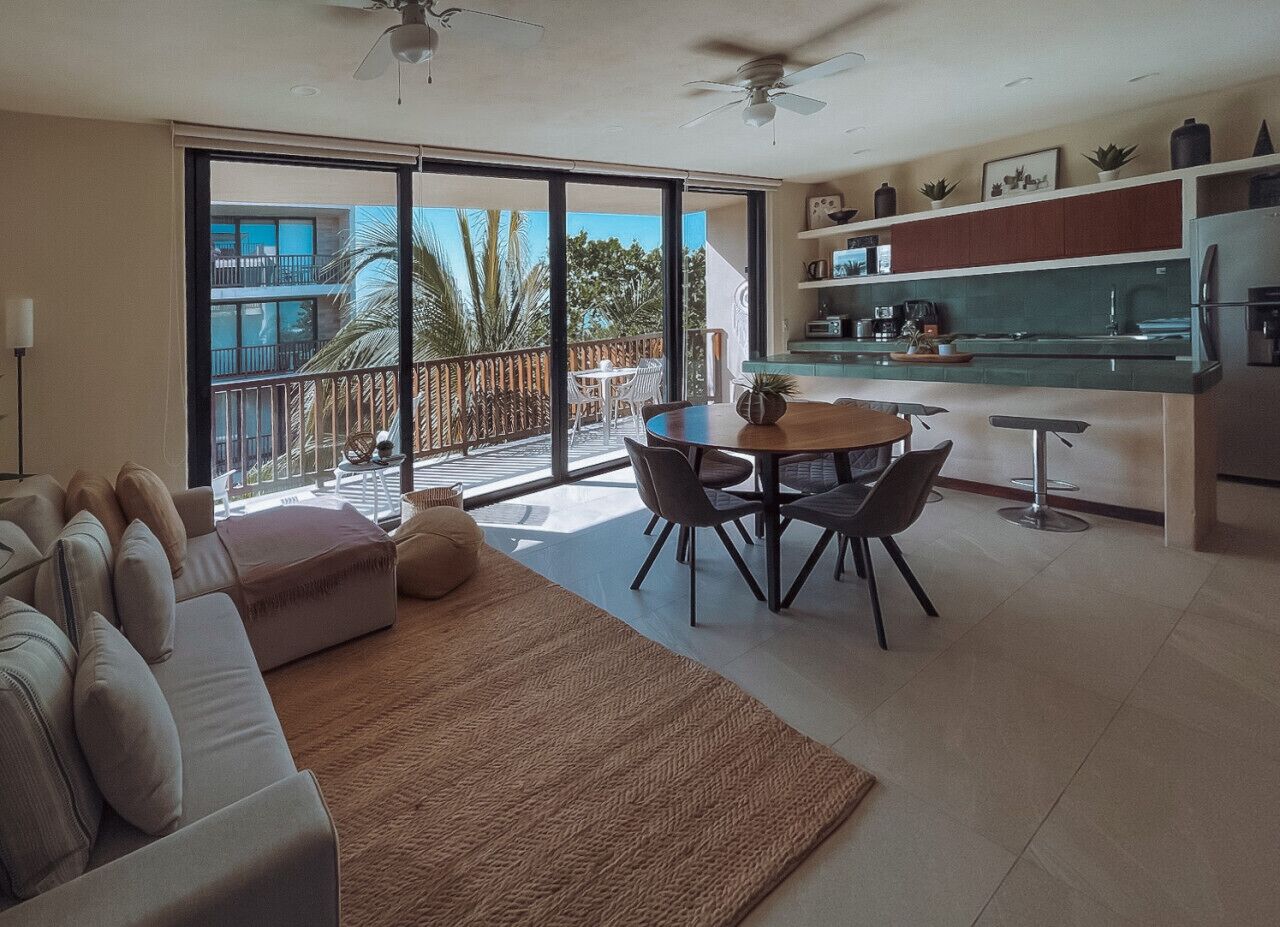 A modern living room with a sofa, a coffee table, and a dining area with chairs. There is a balcony with a view of palm trees and a building.