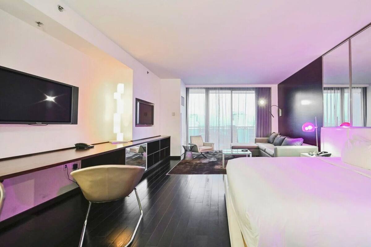 A modern hotel room with a large bed, a desk with a flat-screen TV, a sofa, and a large window offering a city view.