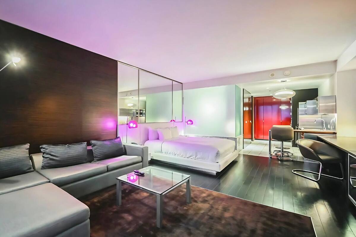 A modern hotel room with a sofa, a glass coffee table, a bed, a desk, and a chair.