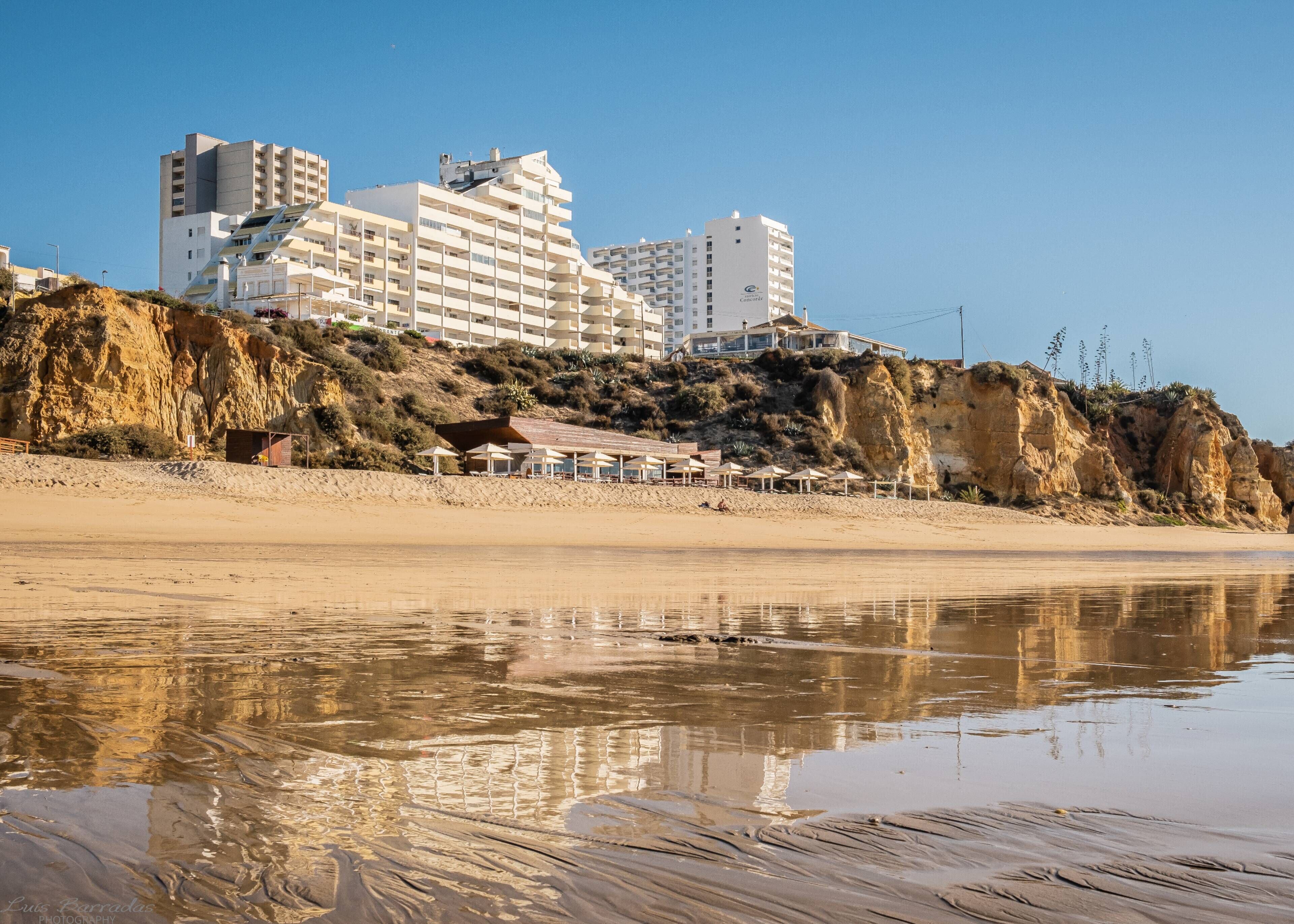A coastal hotel with multiple floors, a sandy beach in the foreground, and rocky cliffs.