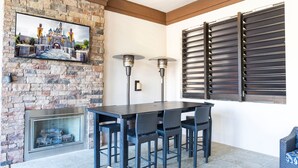 A stone fireplace, a dining table with chairs, a TV mounted on the wall, and a window with blinds. - Solterra 6BR | Private Pool & 4 Ensuites (davenport)
