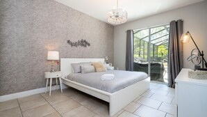 A modern bedroom with a large bed, a nightstand, a lamp, and a view of the outdoors through a glass door. - Solterra 6BR | Private Pool & 4 Ensuites (davenport)