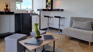 Un moderno salón con un sofá gris, una mesa de centro de madera con libros, un televisor negro y una zona de cocina con un refrigerador negro, taburetes de bar y un ventanal con vistas.