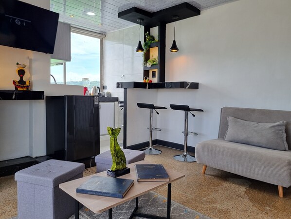Un moderno salón con un sofá gris, una mesa de centro de madera con libros, un televisor negro y una zona de cocina con un refrigerador negro, taburetes de bar y un ventanal con vistas.