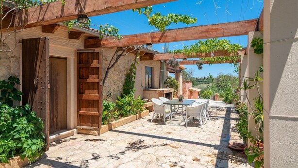 Eine Steinterrasse mit Tisch und Stühlen, eine Holztür und eine mit Weinreben bewachsene Pergola.