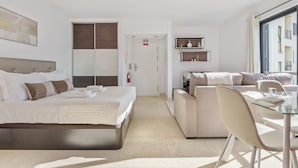 Moderni hotellihuone, jossa on suuri sänky, sohva, lasinen ruokapöytä ja pieni keittiöalue.
