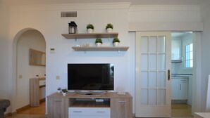 Een woonkamer met een televisie op een houten standaard, een open haard en een deur die naar de keuken leidt.