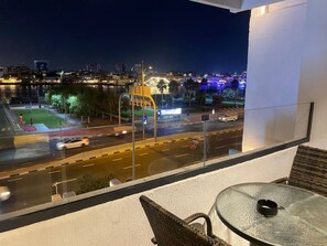 A balcony with a glass table and chairs, overlooking a city street at night with illuminated buildings and a clear view of the road. - LOOKING GOOD 5 MIN 2BHK (Dubai)