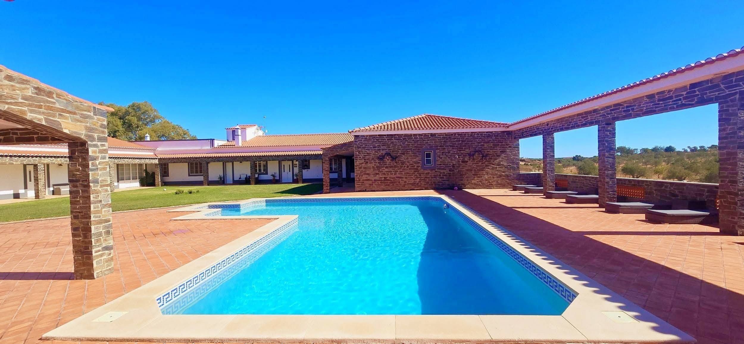Una piscina con un motivo a rilievo geometrico, circondata da una terrazza con pilastri in pietra e un'area salotto coperta.