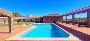 Ein Schwimmbecken mit geometrischem Fliesenmuster, umgeben von einer Terrasse mit Steinpfeilern und einer überdachten Sitzgelegenheit.
