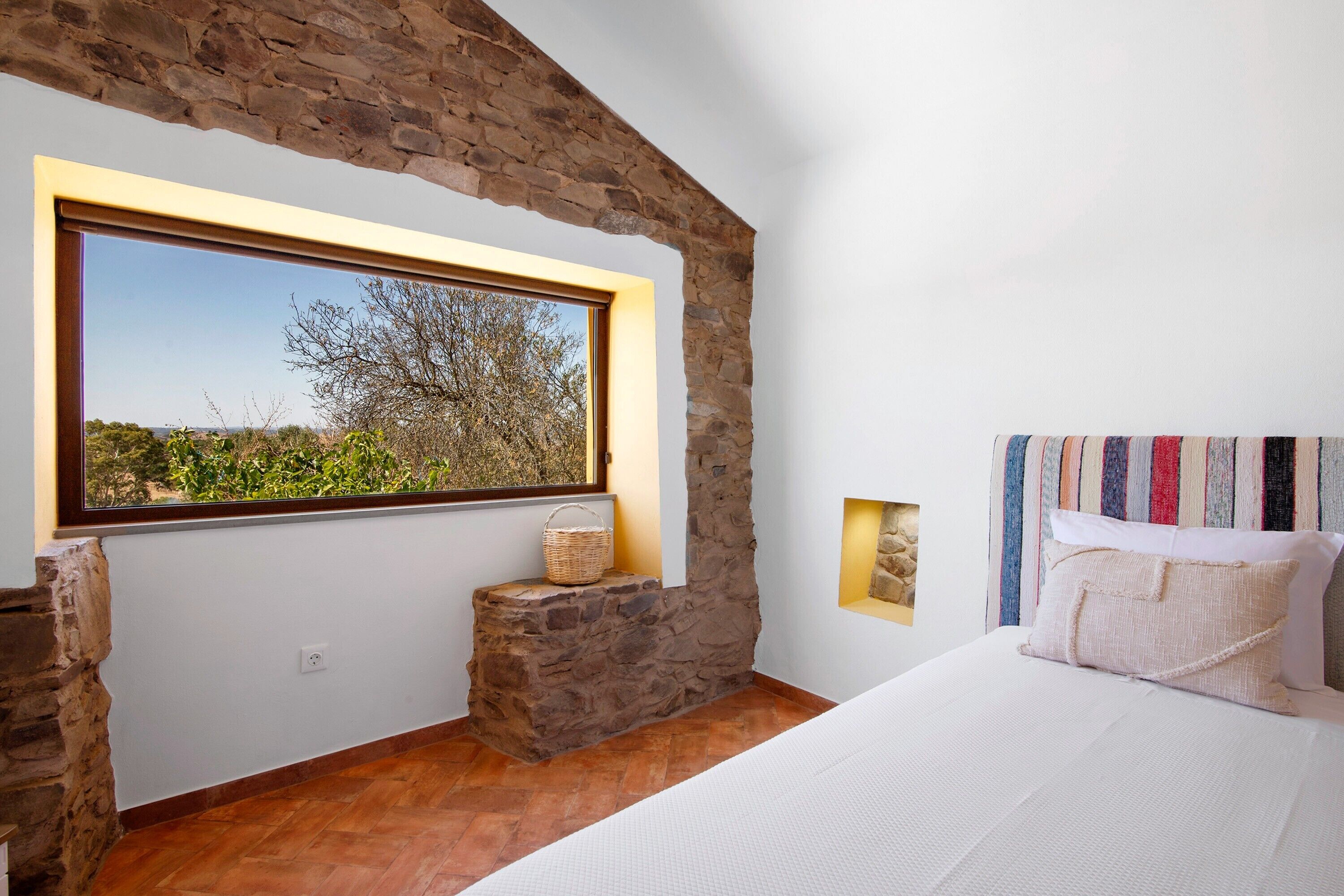 Une chambre avec un mur en pierre, une grande fenêtre offrant une belle vue, un lit avec une literie blanche et une tête de lit à rayures.