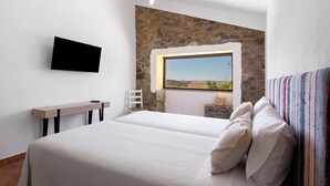 A bedroom with a stone wall, a large window, a bed with white bedding, a bedside table, and a mounted TV.