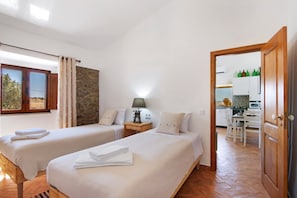 Un dormitorio con dos camas, una pared de piedra, suelo de madera y una cocina visible a través de una puerta abierta.