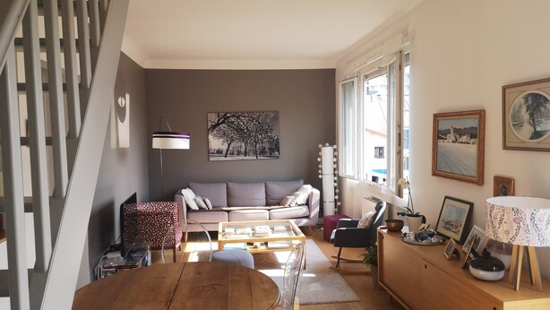 A living room with a sofa, a coffee table, and a dining table. There are windows, a lamp, and a painting on the wall.