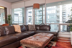Ein modernes Wohnzimmer mit einem ledernen Ecksofa, einem gepolsterten Hocker und großen Fenstern mit Blick auf einen Wolkenkratzer.