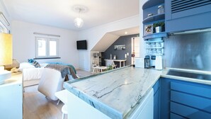 A modern kitchen with a marble countertop, blue cabinets, and a built-in stove. There is a dining area with a table and chairs, and a bedroom with a bed and a window.