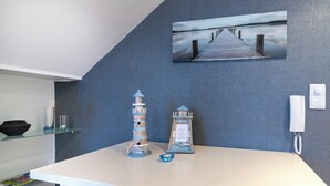 A room with a white table, a blue wall, a painting of a pier, a lighthouse model, a small bowl, and a telephone.