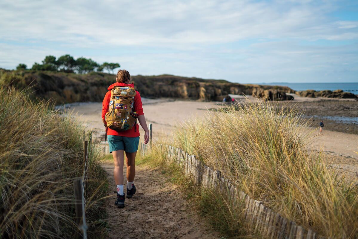 En person går på en sandstig med högt gräs på båda sidor, och stigen leder mot en strand.