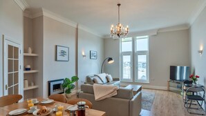A modern living room with a dining area, a sofa, a TV, and a chandelier. - Apartment in Cornwall with Towan Beach Views (Newquay)