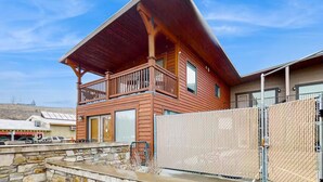 A two-story wooden cabin with a balcony, a stone foundation, and a fenced yard.