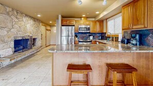 A modern kitchen with a stone fireplace, wooden cabinets, a stainless steel refrigerator, and a large island with stools.