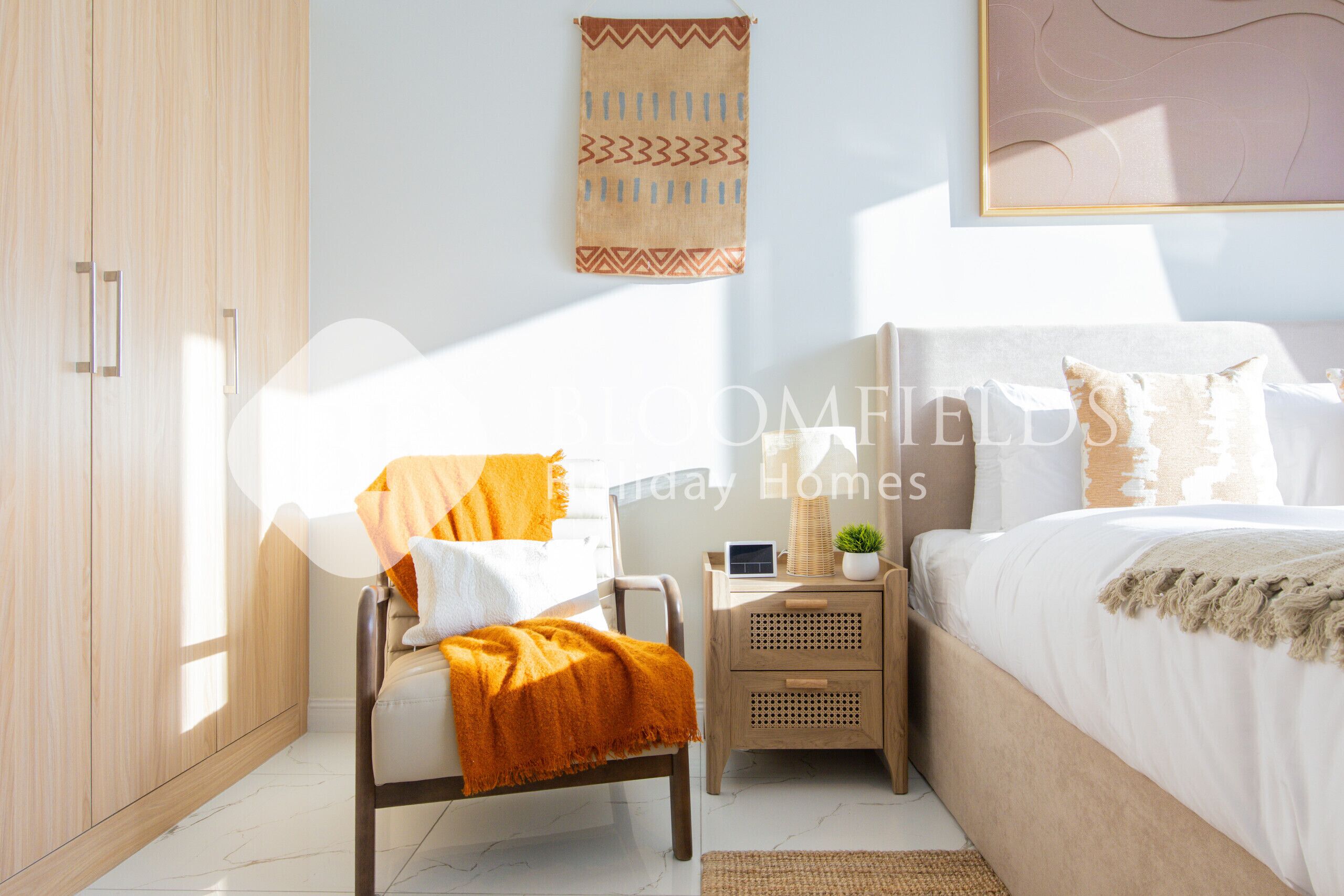 Un dormitorio con cama, una silla con manta, una mesita de noche con lámpara y un tapiz colgado en la pared.