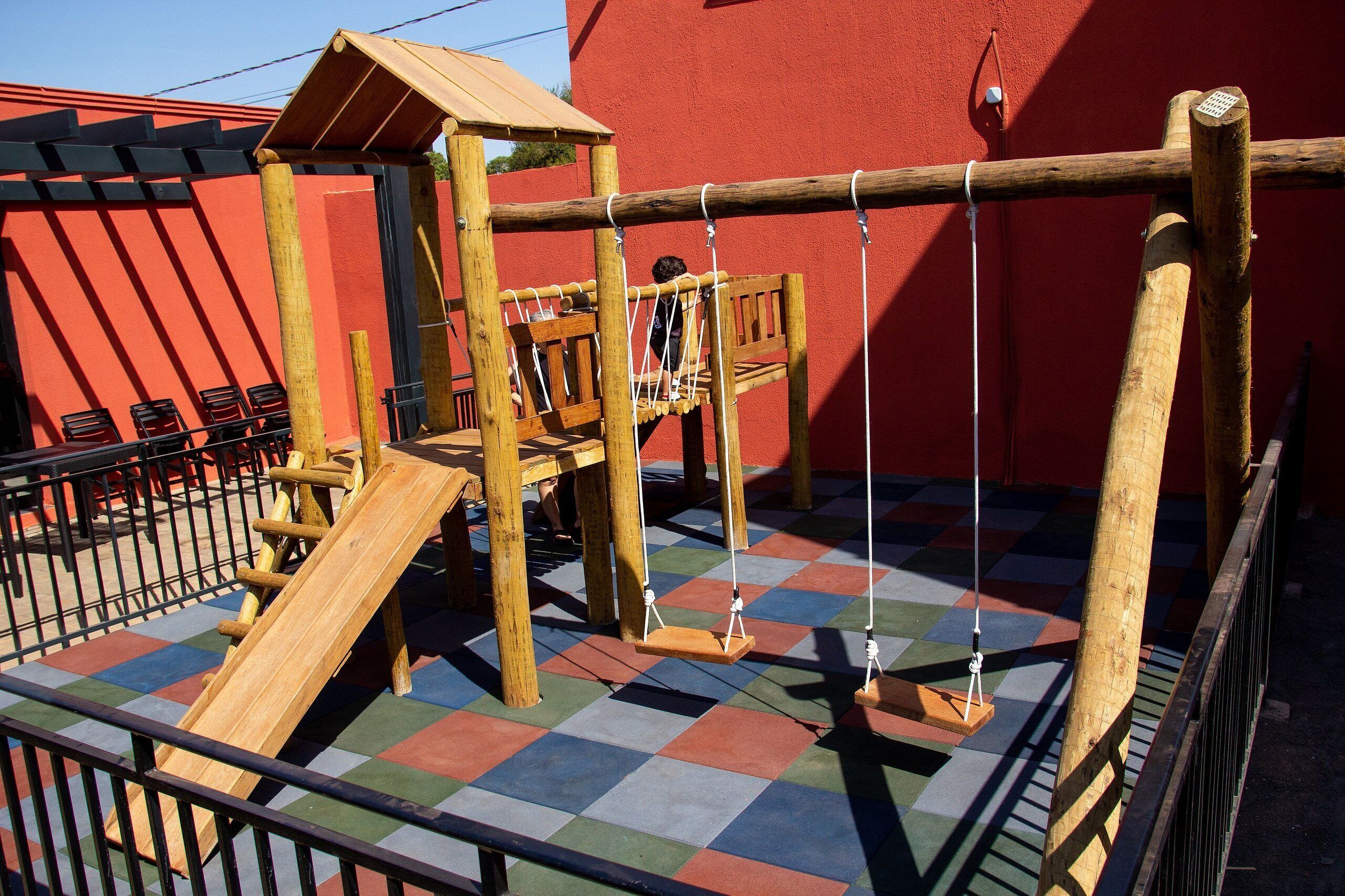 Una estructura de juego de madera con un tobogán y columpios, frente a un muro rojo.
