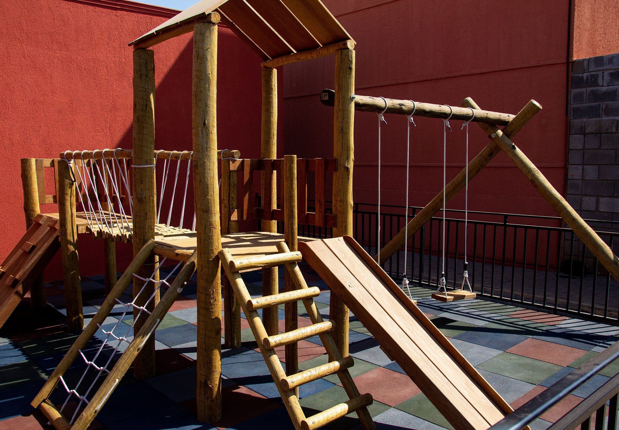 A wooden play structure with a ladder, slide, and rope bridge against a red wall.