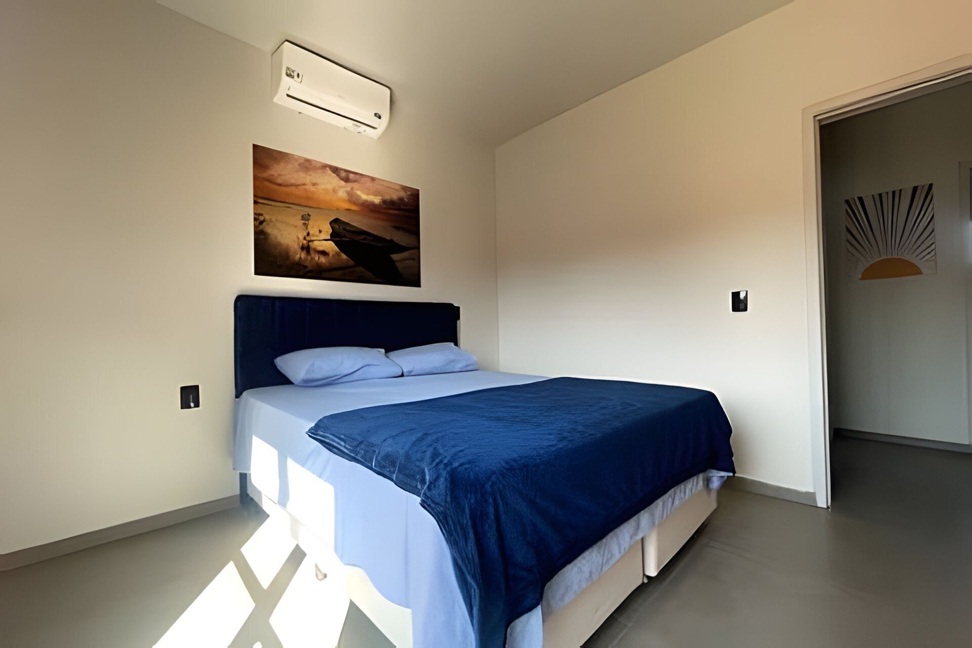 A bedroom with a bed, a painting, an air conditioning unit, and a door with a sun-like design.