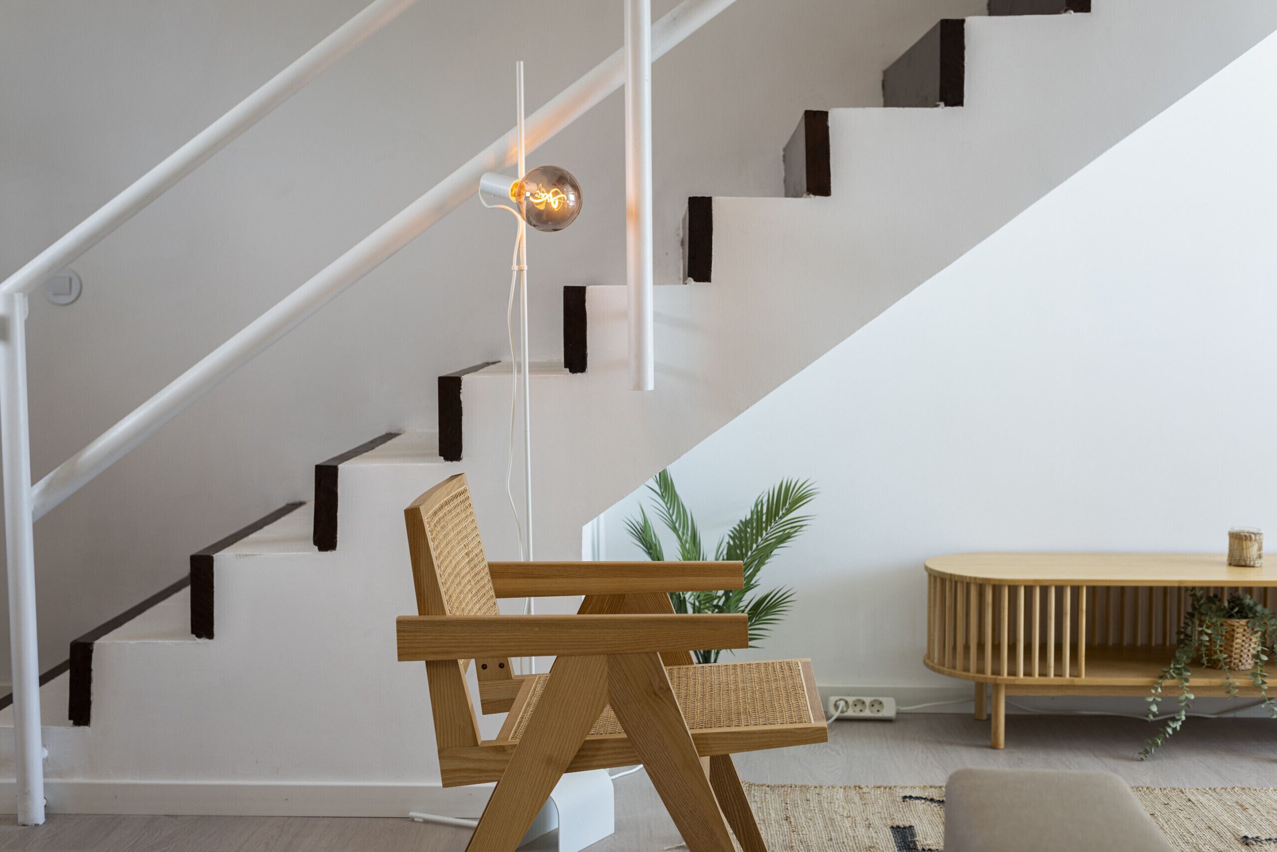 Eine Treppe mit weißen Wänden und schwarzen Akzenten, ein Holzstuhl, ein kleiner Tisch mit einer Pflanze und eine hängende Leuchte.