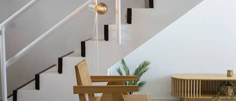 Una escalera con paredes blancas y detalles en negro, una silla de madera, una pequeña mesa con una planta y una lámpara colgante.