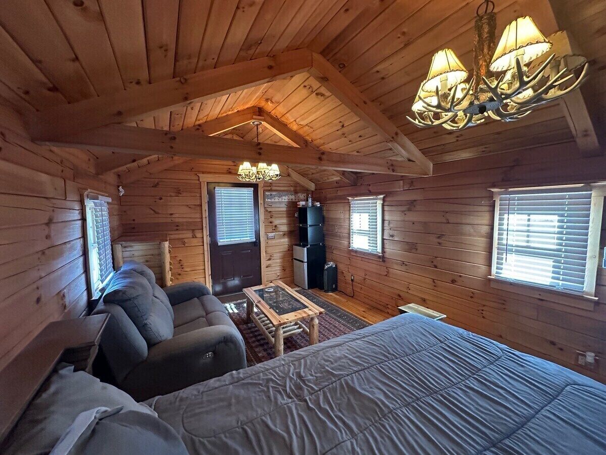 A cozy cabin interior with a large bed, a sofa, a small table, and a chandelier.
