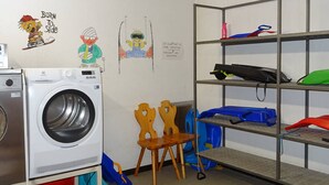 A laundry room with a front-loading washing machine, wooden chairs, a shelf with various items, and children's drawings on the wall.