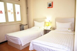 Habitación de hotel con dos camas individuales, una mesita de noche con lámpara, un radiador y una ventana con cortinas.