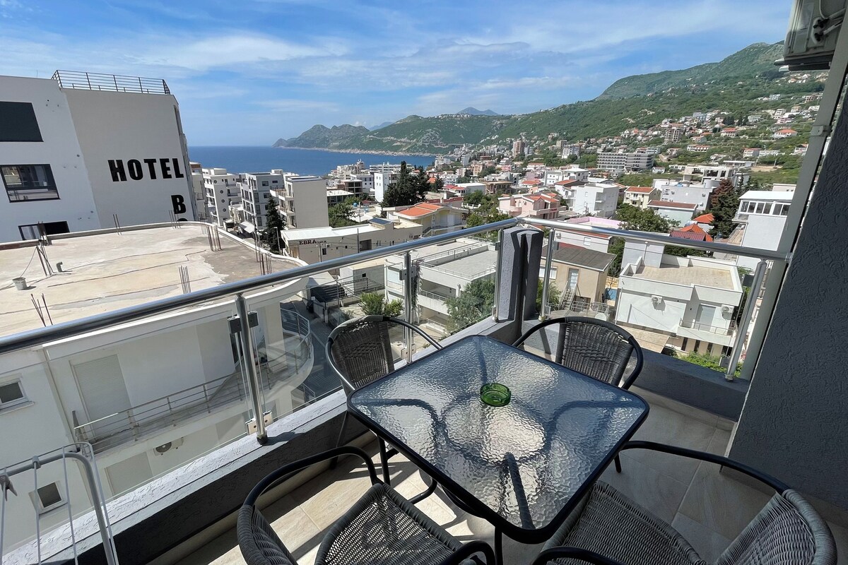 Un balcón con una mesa y sillas de vidrio, con vistas a un pueblo costero y al mar.