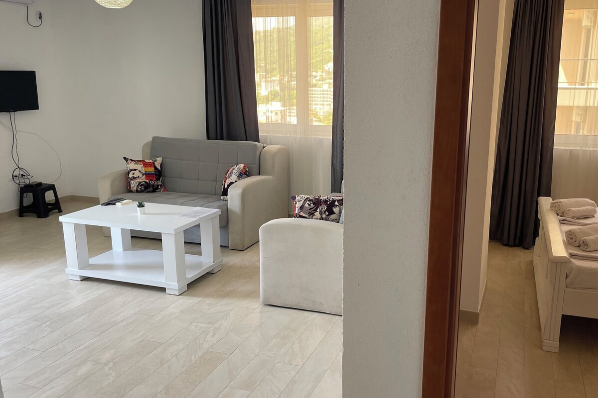 A modern living room with a grey sofa, a white coffee table, and a bed with neatly folded towels.