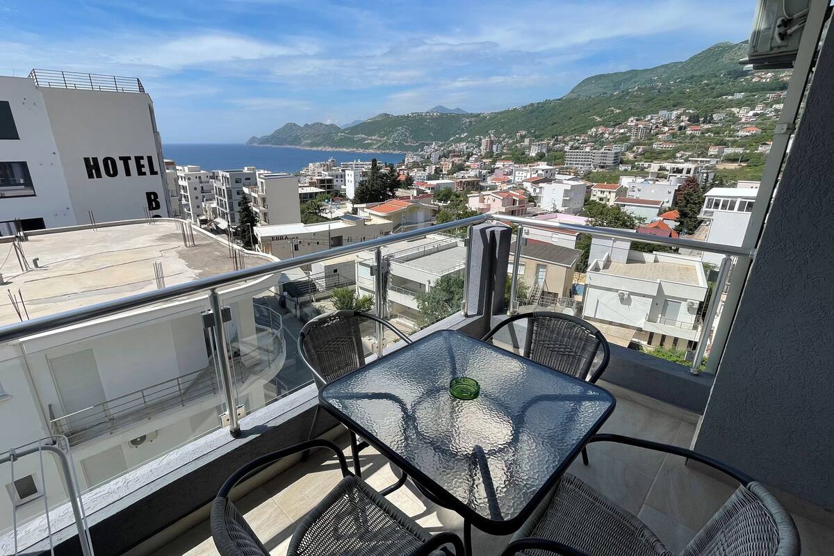 A balcony with a glass table and chairs, overlooking a coastal town with buildings and a view of the sea.